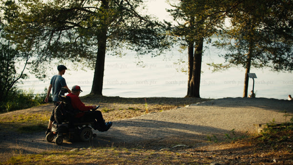 Young man pushes senior man in wheelchair through summer park by lake