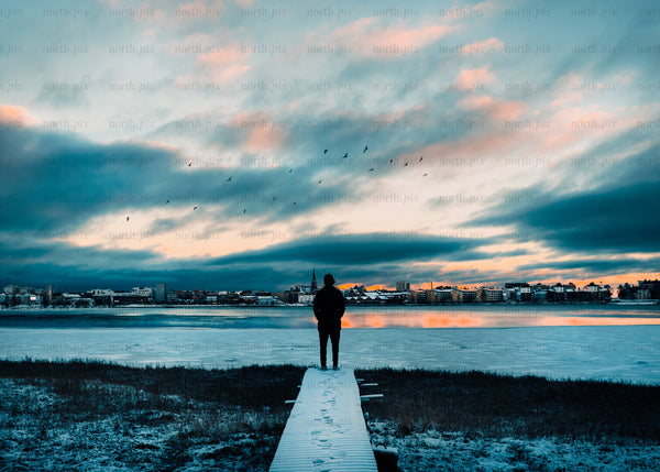 Solitary figure overlooks frozen lake by cityscape at sunset