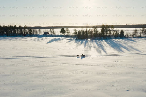 Snowy frozen lake scene with long tree shadows and snowmobile riders in winter light