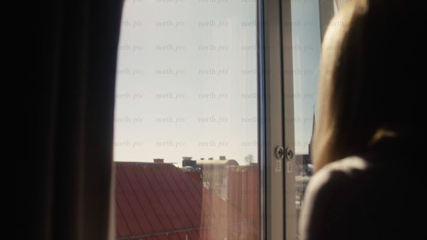 Woman looking out hotel window at sunny cityscape in Luleå