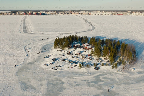 Snowy island village on a frozen lake with trees, cabins, and city skyline in background