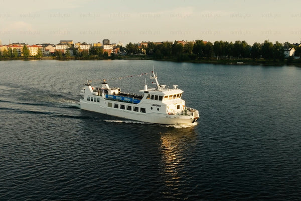 White passenger ferry gliding across calm lake near Luleå city center with trees and buildings