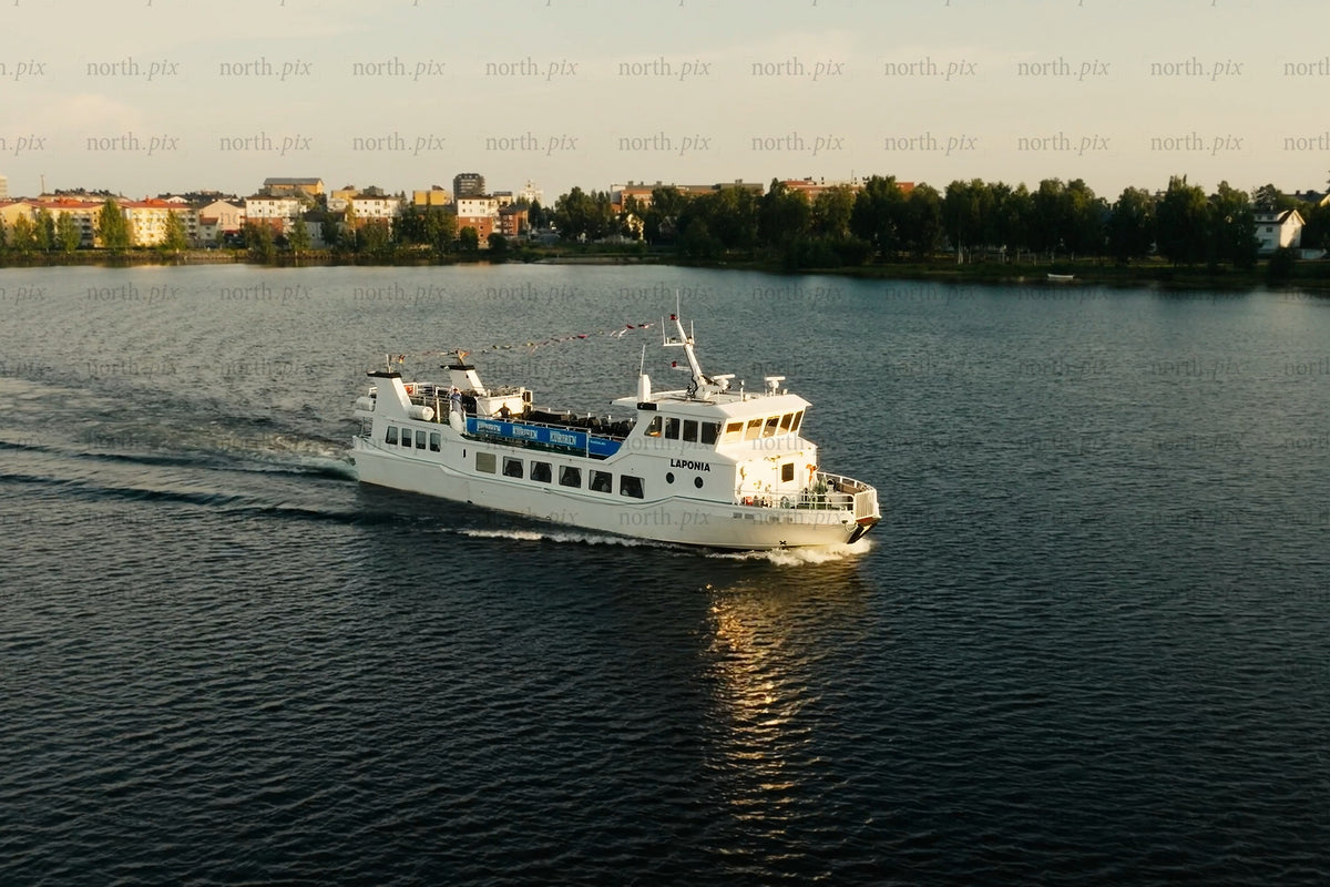 White passenger ferry gliding across calm lake near Luleå city center with trees and buildings