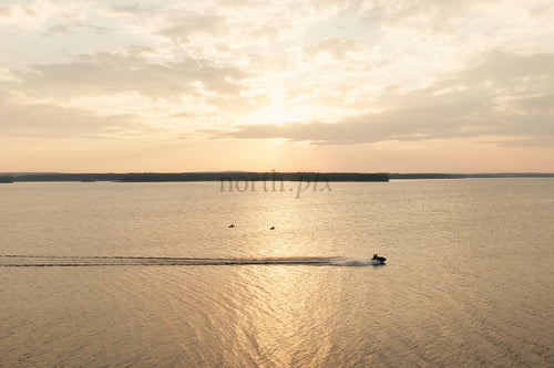 Gyllene solnedgång över lugn sjö med båtvågor och avlägsen östrandlinje vid Luleå centrum