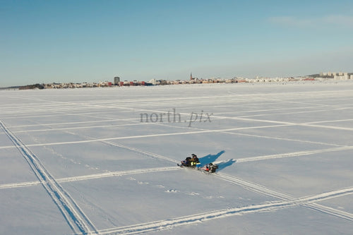 Snöig frusen sjö med stadssilhuett och snöskotrar under klarblå himmel