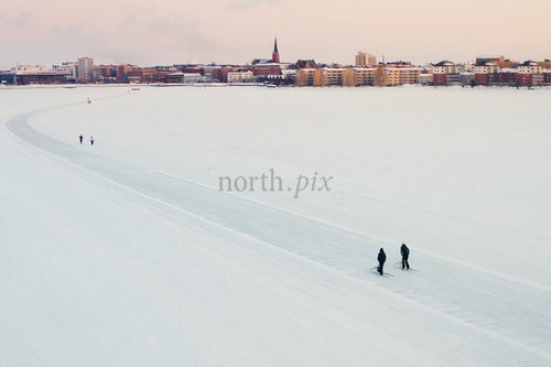 Snöig sjö i förgrunden med Luleå stadssilhuett under vinterns lugna, mjuka solnedgång