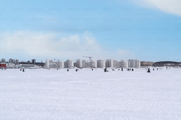 People ice fishing on frozen bay with waterfront apartments, winter