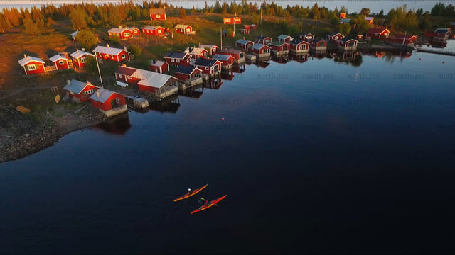 Kayakers paddle past red cabins in the Luleå archipelago at sunset
