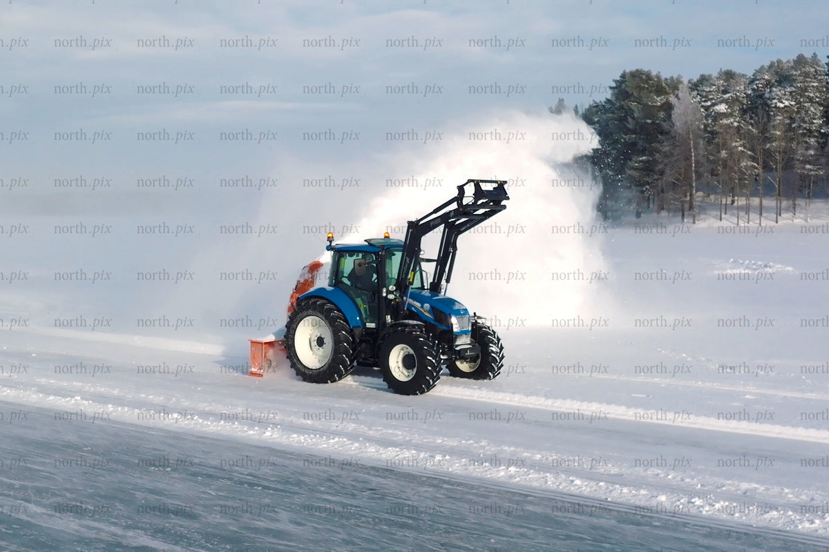 Blue tractor with snow plow clearing snow in open field under winter sky near forest