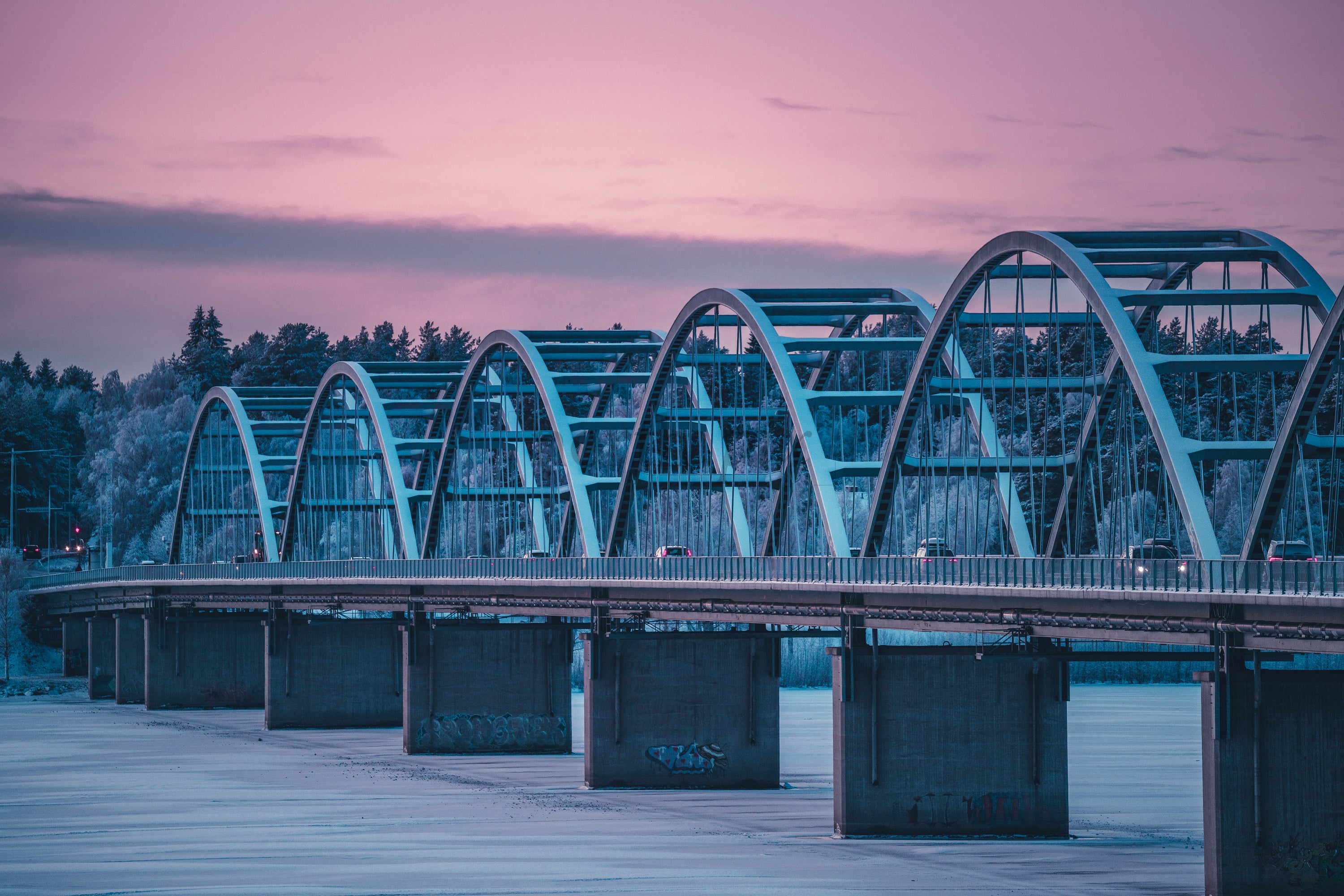 Arched Steel Bridge Over Frozen River At Pink Sunset With Forested Shoreline And Evening Traffic