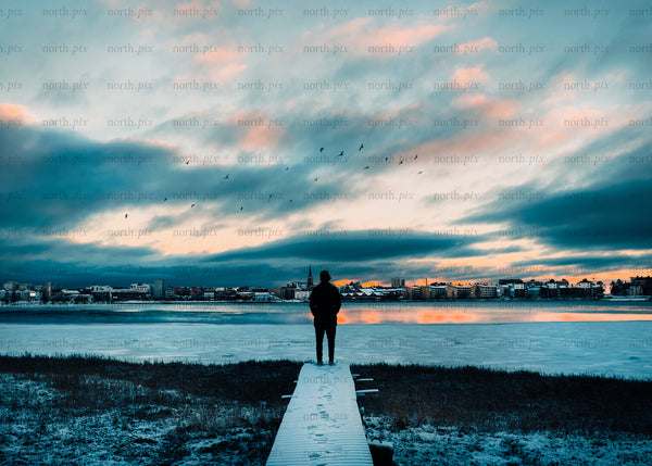 Solitary figure overlooks frozen lake by cityscape at sunset