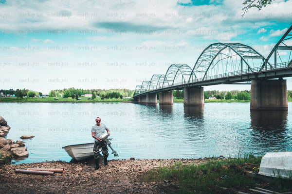 Man with outboard motor on riverbank under arched bridge, summer