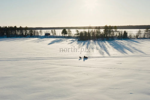Snöig frusen sjöscen med långa trädskuggor och snöskoteråkare i vinterljus
