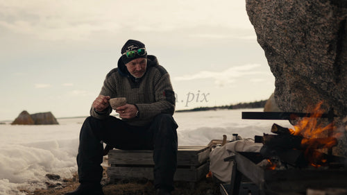 Man njuter av varm dryck vid brasa på snöig strand i Luleå skärgård, vinter