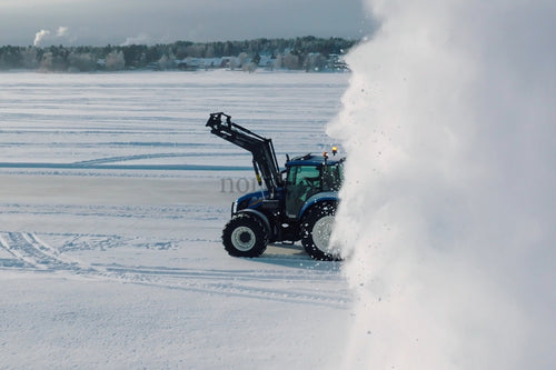 Blå traktor röjer snö i öppet fält med stort snömoln i vinterlandskap