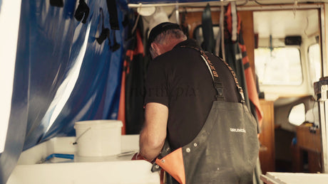 Fisherman Cleans Fish on Boat While Wearing Orange Gloves and Overalls