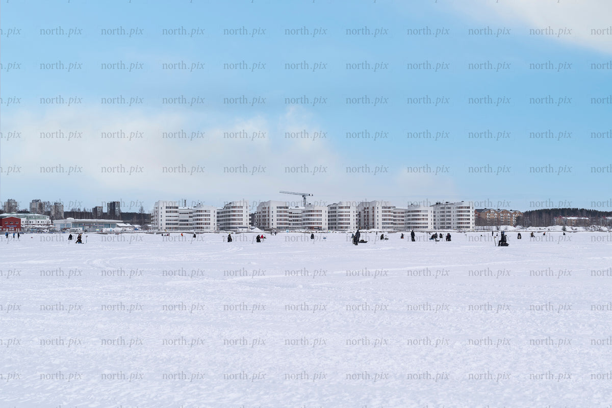 People ice fishing on frozen bay with waterfront apartments, winter