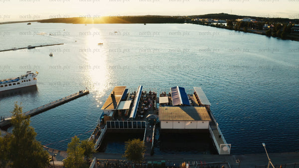 Luleå: Sunset over a floating restaurant on a summer evening. Aerial view of people dining, boats sailing, and a scenic waterfront.