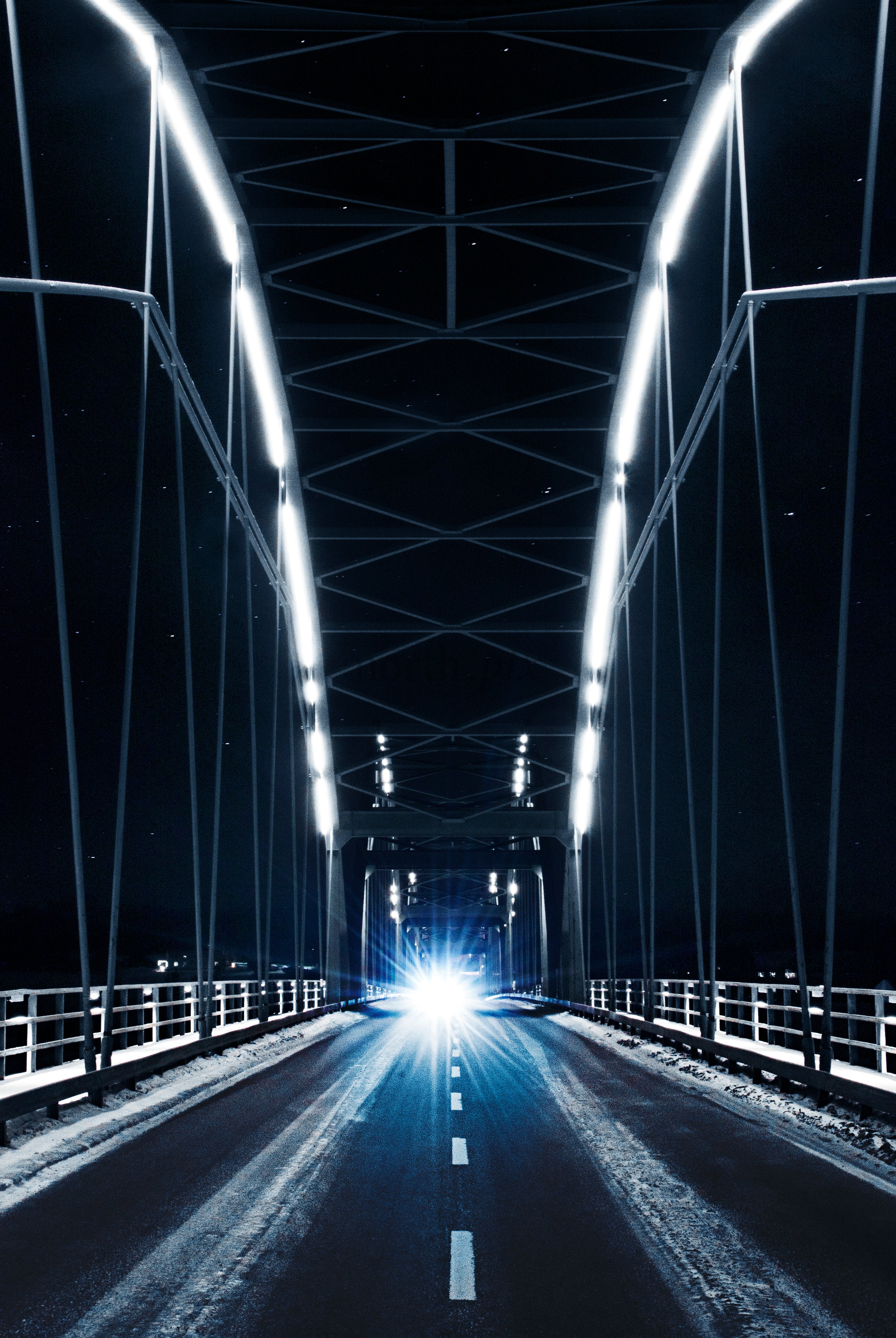 Car with bright headlights crossing a lit truss bridge at night