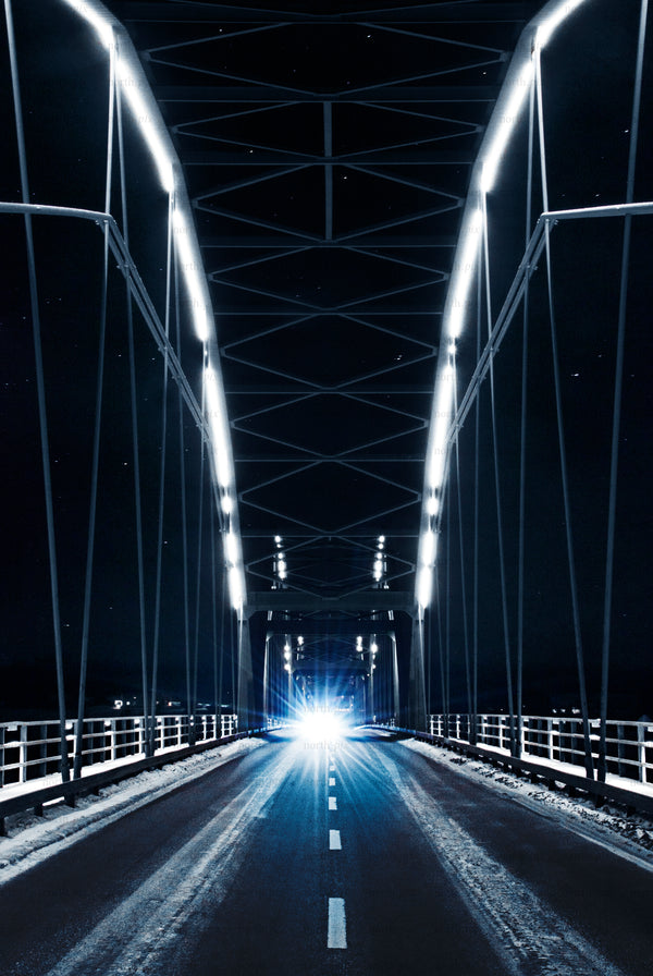Car with bright headlights crossing a lit truss bridge at night