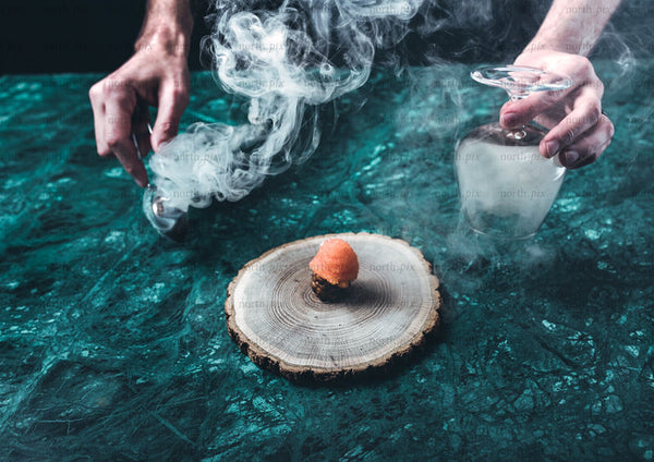 Hands lifting glass cloche over smoked appetizer on wood
