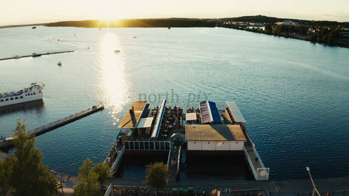 Luleå, Sverige: Solnedgång över en flytande restaurang en sommarkväll. Flygfoto över människor som äter, båtar som seglar och en vacker strandpromenad.