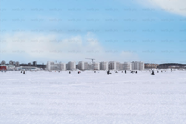 People ice fishing on frozen bay with waterfront apartments, winter