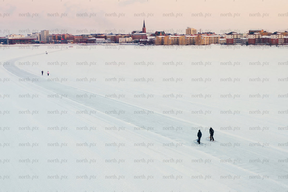 Snöig sjö i förgrunden med Luleå stadssilhuett under vinterns lugna, mjuka solnedgång